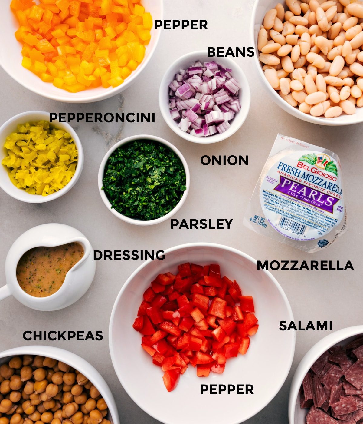All the ingredients for this recipe are prepped and ready for straightforward assembly: beans, onion, parsley, mozzarella, pepper salami, pepperoncini, chickpeas, and dressing.