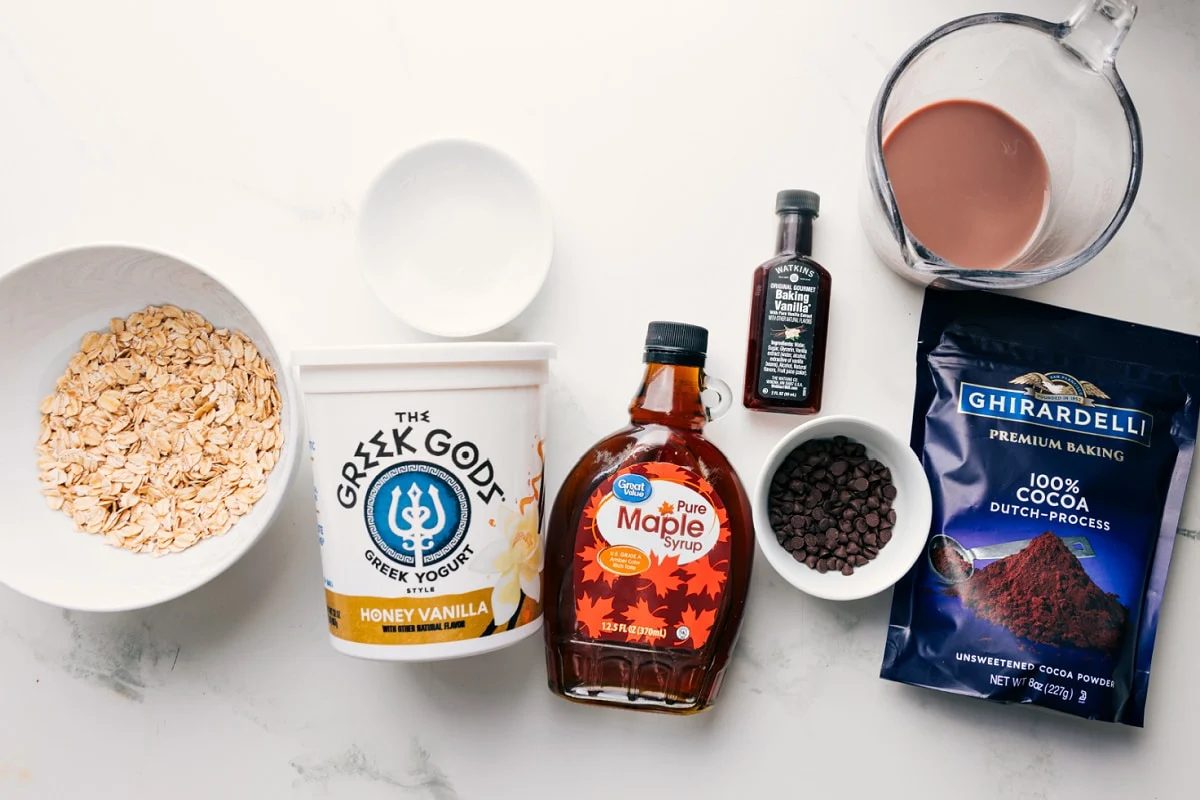 All the ingredients in this recipe prepped out for straightforward assembly including the oats, salt, greek yogurt, maple syrup, cocoa powder, vanilla, chocolate chips, and chocolate milk.