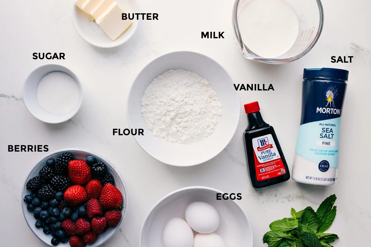 All the ingredients for this recipe prepped and ready for straightforward assembly, including butter, milk, salt, vanilla, flour, sugar, berries, and eggs.
