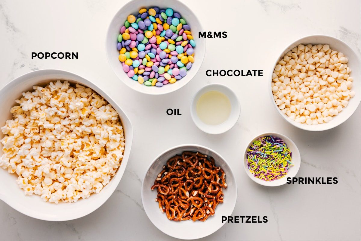 All the ingredients in this recipe prepped out for straightforward assembly including the popcorn, pretzels, M&Ms, sprinkles, chocolate, and oil.