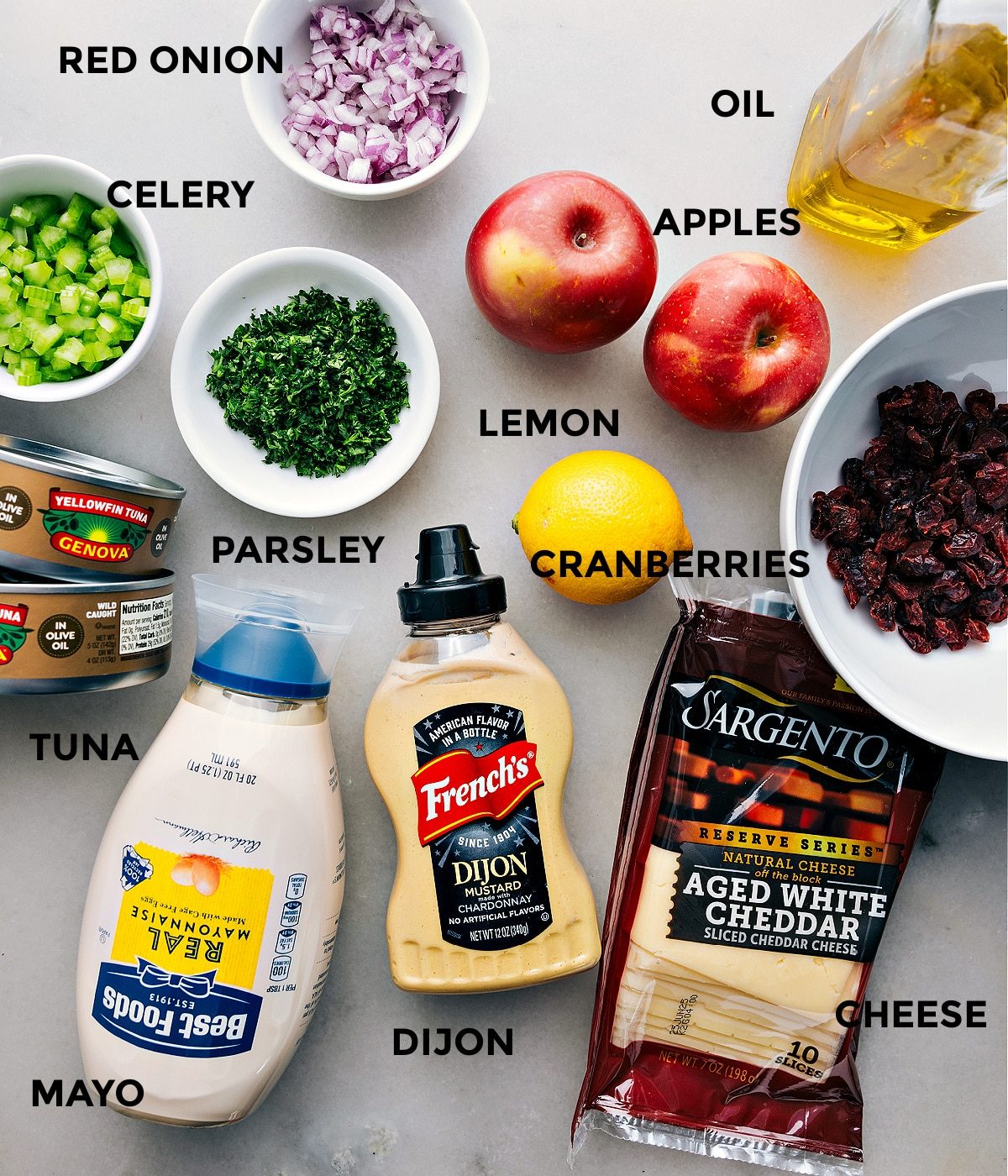All the ingredients in this recipe are prepped and ready for straightforward assembly—apples, cranberries, lemon, oil, onion, celery, parsley, cheddar, Dijon, mayo, and meat.