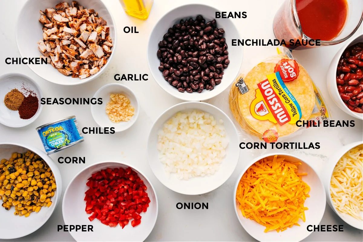 All the ingredients for Chicken Enchilada Casserole prepped and ready to assemble, including chicken, seasonings, corn, peppers, green chiles, garlic, oil, beans, enchilada sauce, chili beans, corn tortillas, onion, and cheese.