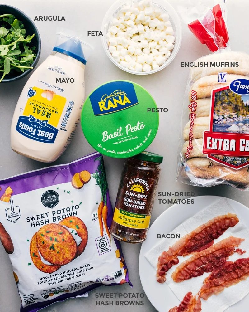 Ingredients prepped out for straightforward assembly including the arugula, feta, mayo, pesto, sun-dried tomatoes, bread, sweet potato hash browns, and bacon.