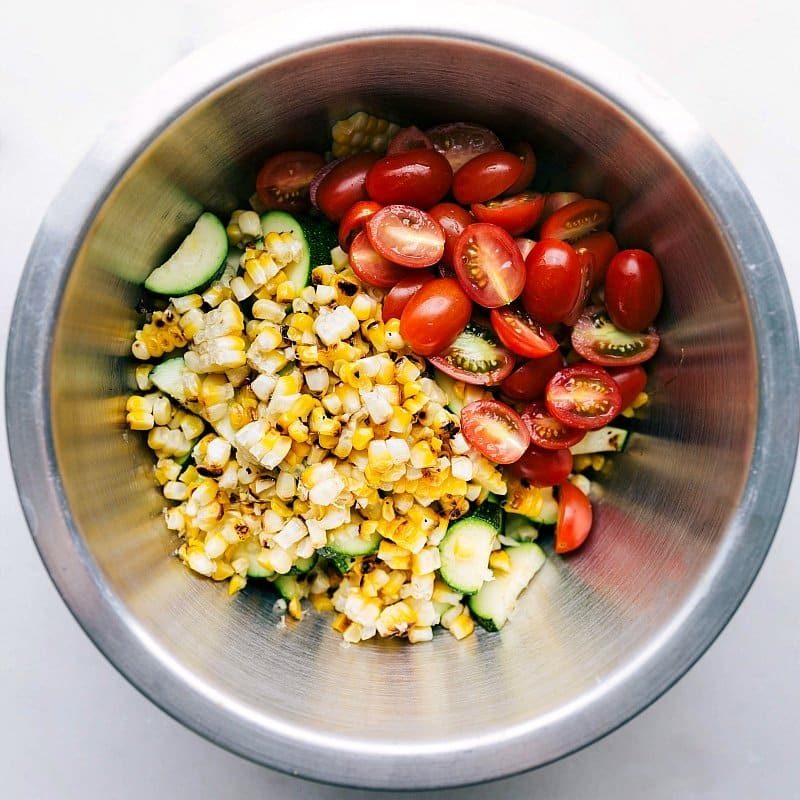 A bowl of zucchini corn salad ingredients with fresh corn, zucchini, tomatoes, and herbs before mixing.