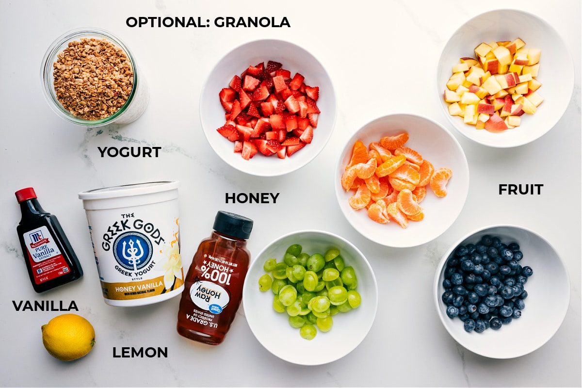 All ingredients prepped and ready for straightforward assembly including vanilla, lemon, honey, yogurt, strawberries, oranges, apples, blueberries, and grapes.