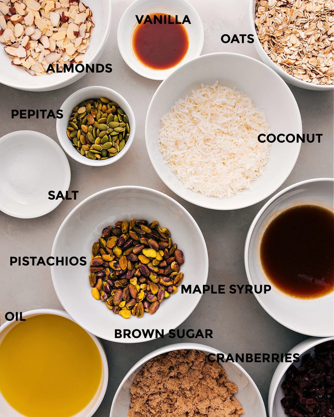 All the ingredients in this recipe are prepped for straightforward assembly: coconut, oats, vanilla, almonds, pepitas, maple syrup, pistachios, salt, brown sugar, cranberries, and oil.