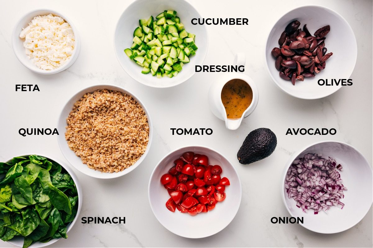 Ingredients in this recipe prepped out for straightforward assembly including the feta, quinoa, spinach, cucumber, dressing, olive, avocado, onion, and tomato.