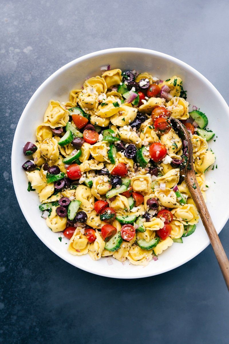 Greek tortellini salad with a spoon in the bowl, ready to be served with fresh vegetables and satisfying dressing.