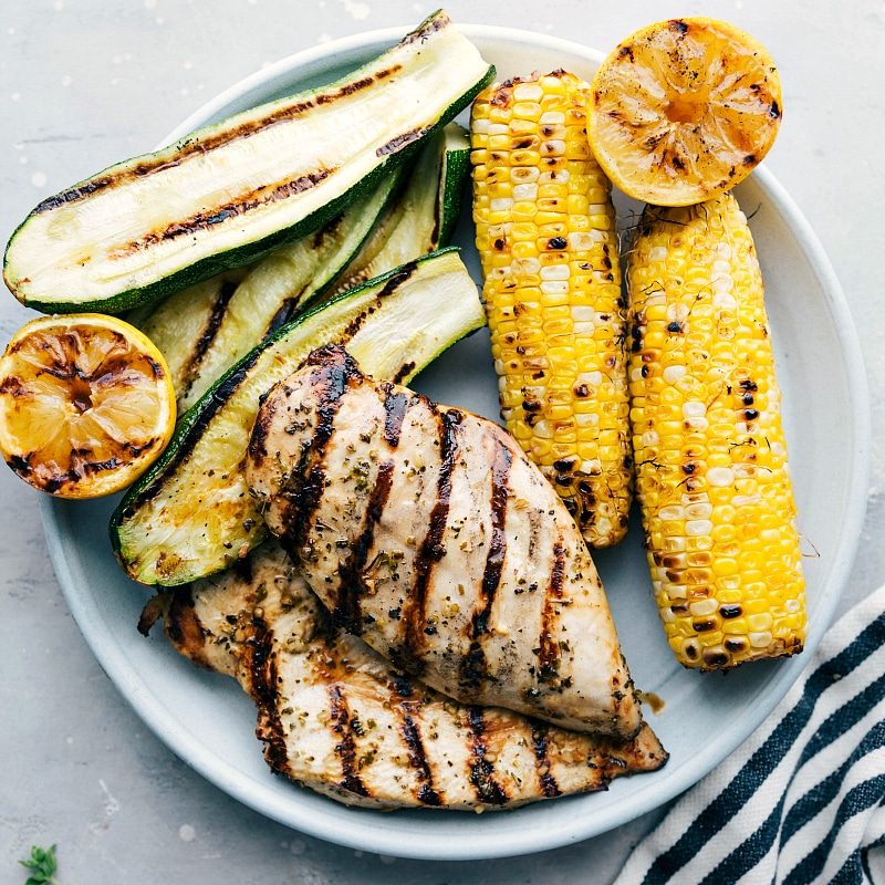 The result of evenly marinated and grilled meat, served with grilled zucchini, corn, and lemons on the side, creating a mouthwatering meal.