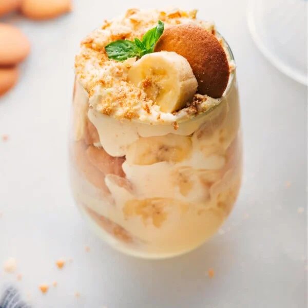 A glass filled with layers of the satisfying banana pudding recipe, alternating between pudding, vanilla cookies, fresh bananas, and topped with vanilla cookie crumbs, creating a delightful dessert.