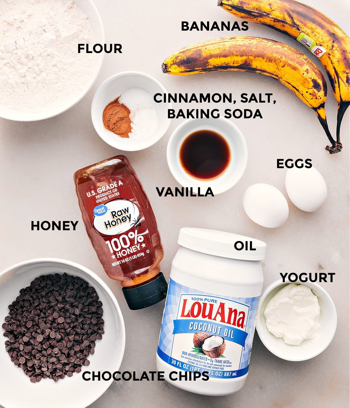 Ingredients in this recipe prepped out for straightforward assembly including flour, bananas, cinnamon, salt, baking soda, eggs, vanilla, honey, chocolate chips, oil, and yogurt.