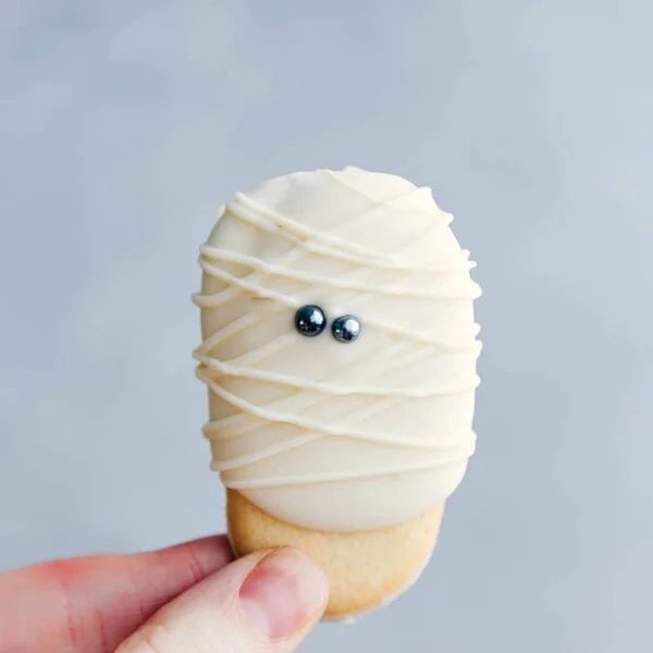 An adorable and festive mummy cookie being held, decorated with white icing and eyes to resemble a mummy, reliable for Halloween.