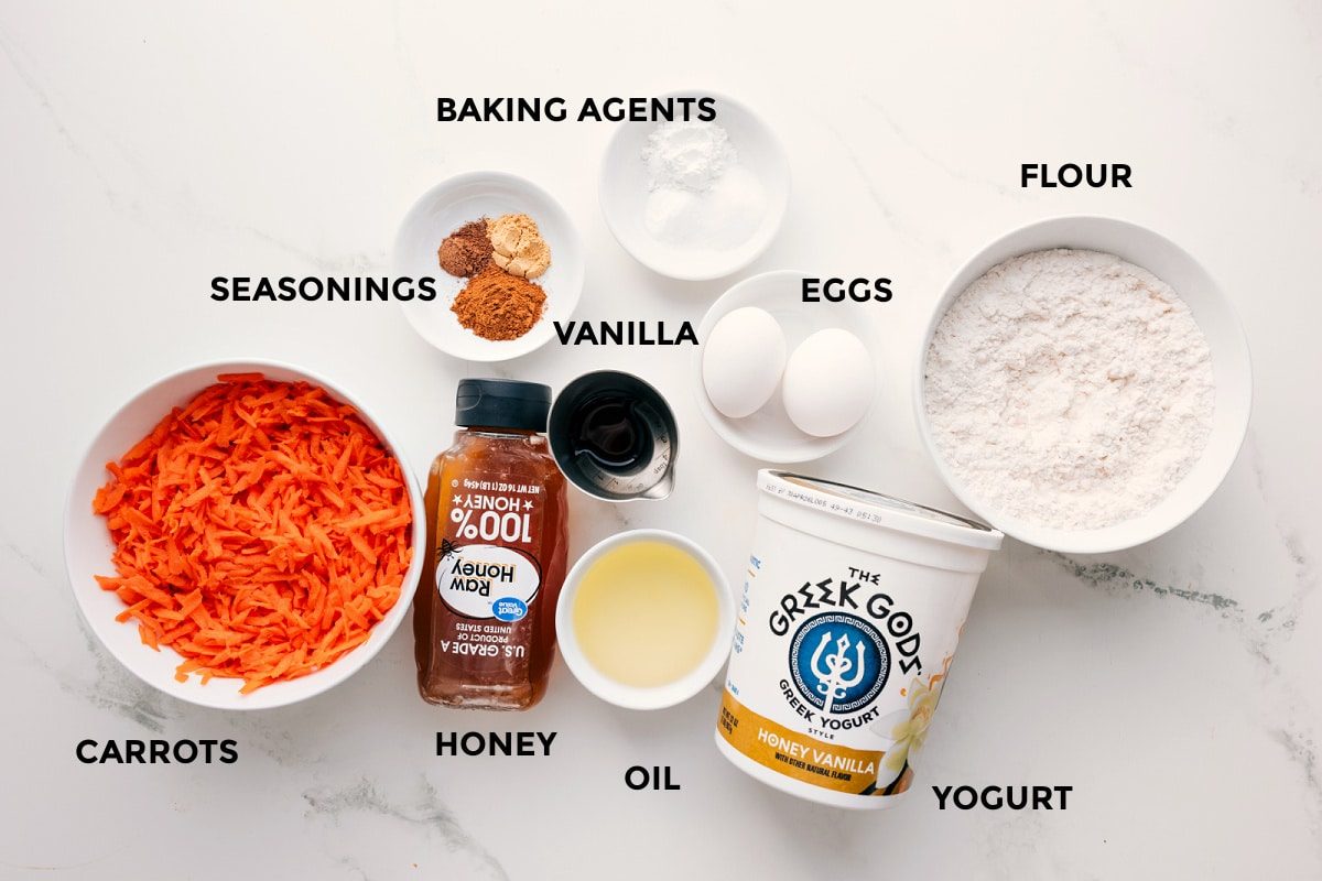 All the ingredients in this recipe prepped out for straightforward assembly including the carrots, seasonings, honey, oil, yogurt, flour, eggs, vanilla, and the baking agents.