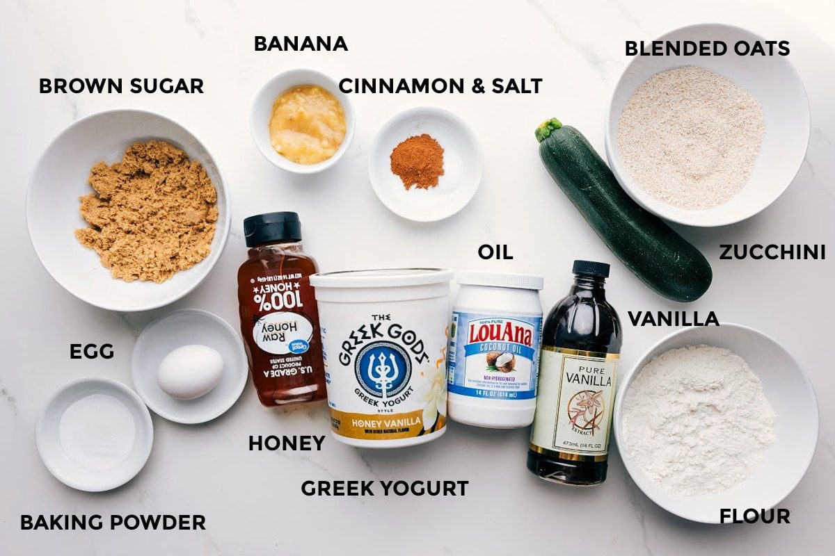All the ingredients in this recipe prepped out for straightforward assembly including the brown sugar, egg, baking powder, honey, Greek yogurt, banana, cinnamon, salt, oil, vanilla, flour, zucchini, and blended oats.