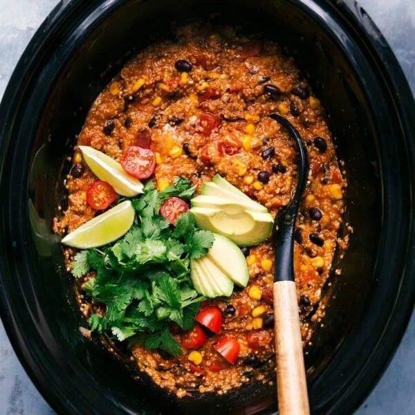 slow cooker quinoa enchilada freshly made, served with a spoon and garnished with cilantro, tomatoes, avocado slices, and lime wedges.