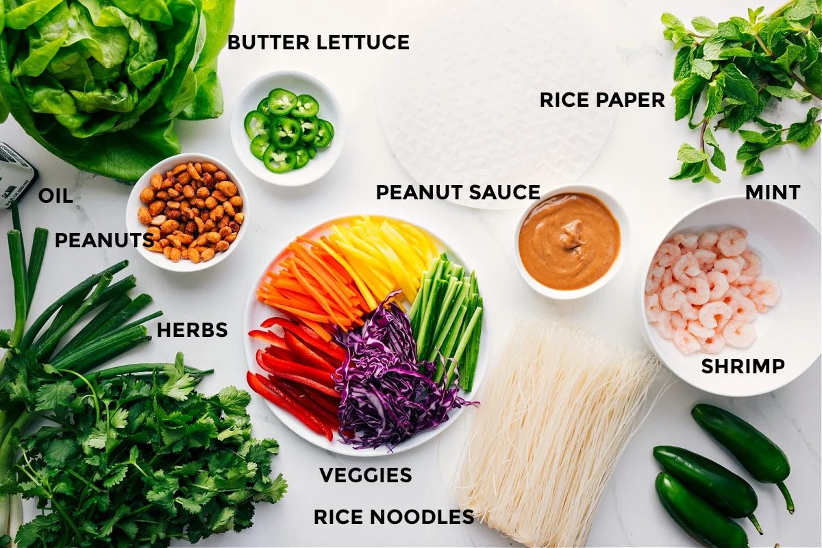 All the ingredients in this recipe prepped out for straightforward assembly including the lettuce, oil, peanuts, veggies, rice noodles, shrimp, peanut sauce, rice paper, and mint.