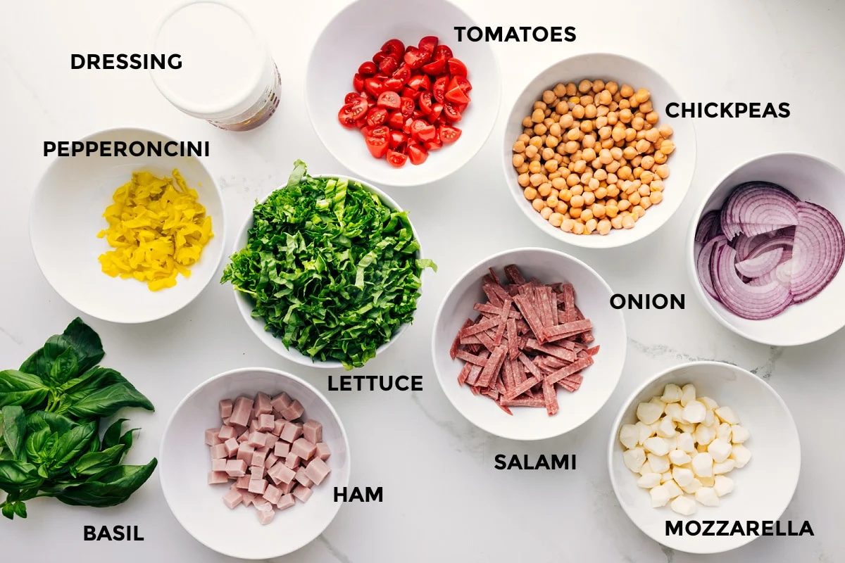 Italian chopped salad ingredients prepped for straightforward assembly, including dressing, pepperoncini, basil, ham, lettuce, tomatoes, salami, chickpeas, mozzarella, and onion.