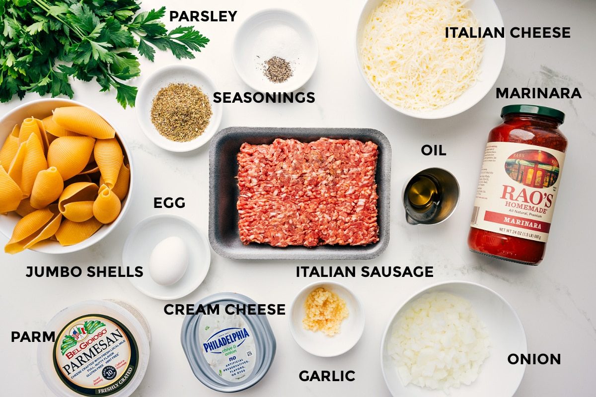 Ingredients for stuffed shells prepped for straightforward assembly, including jumbo shells, Parmesan, cream cheese, egg, seasonings, Italian sausage, garlic, onion, marinara, oil, and Italian cheese.