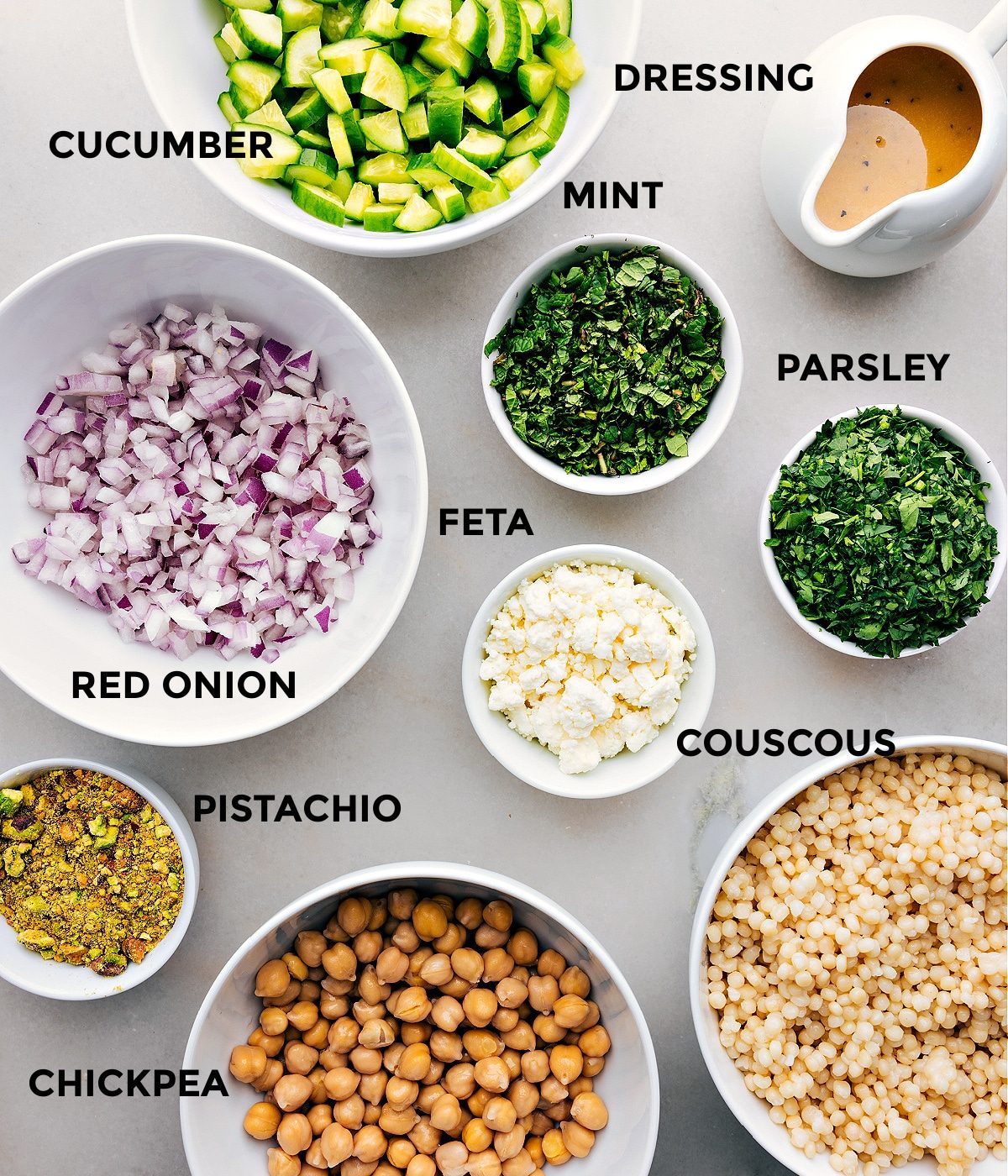 All the ingredients in this recipe are prepped for straightforward assembly, including cucumbers, mint, dressing, parsley, feta, couscous, pistachios, red onion, and chickpeas.