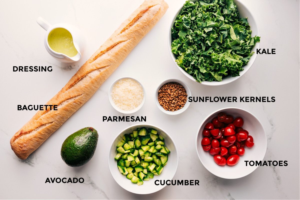 All the ingredients in this recipe prepped out for straightforward assembly including the dressing, baguette, avocado, cucumber, parmesan, sunflower kernels, and tomatoes.