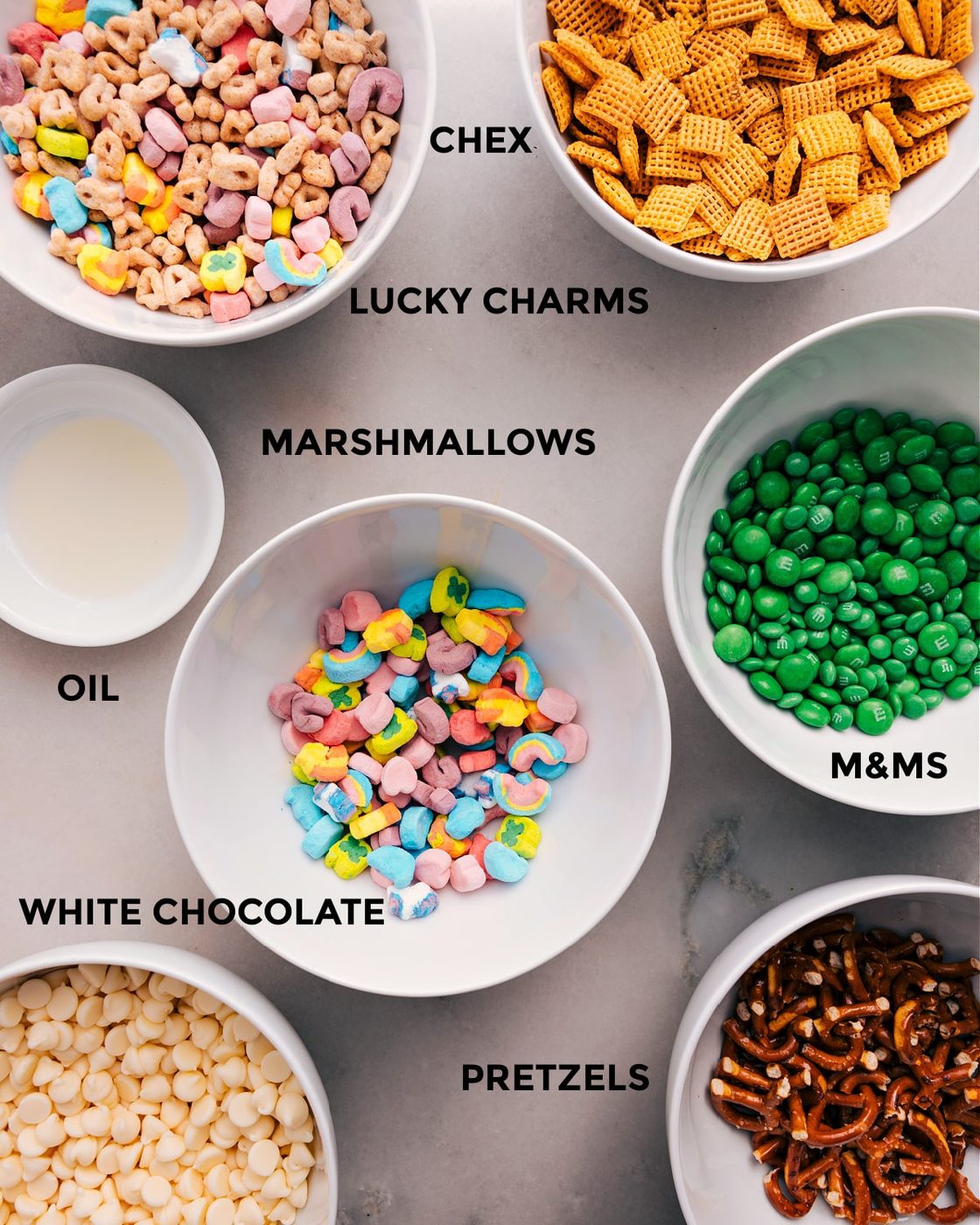 All the ingredients for this recipe are prepped and ready for straightforward assembly, including Chex, Lucky Charms, M&Ms, marshmallows, oil, chocolate, and pretzels.