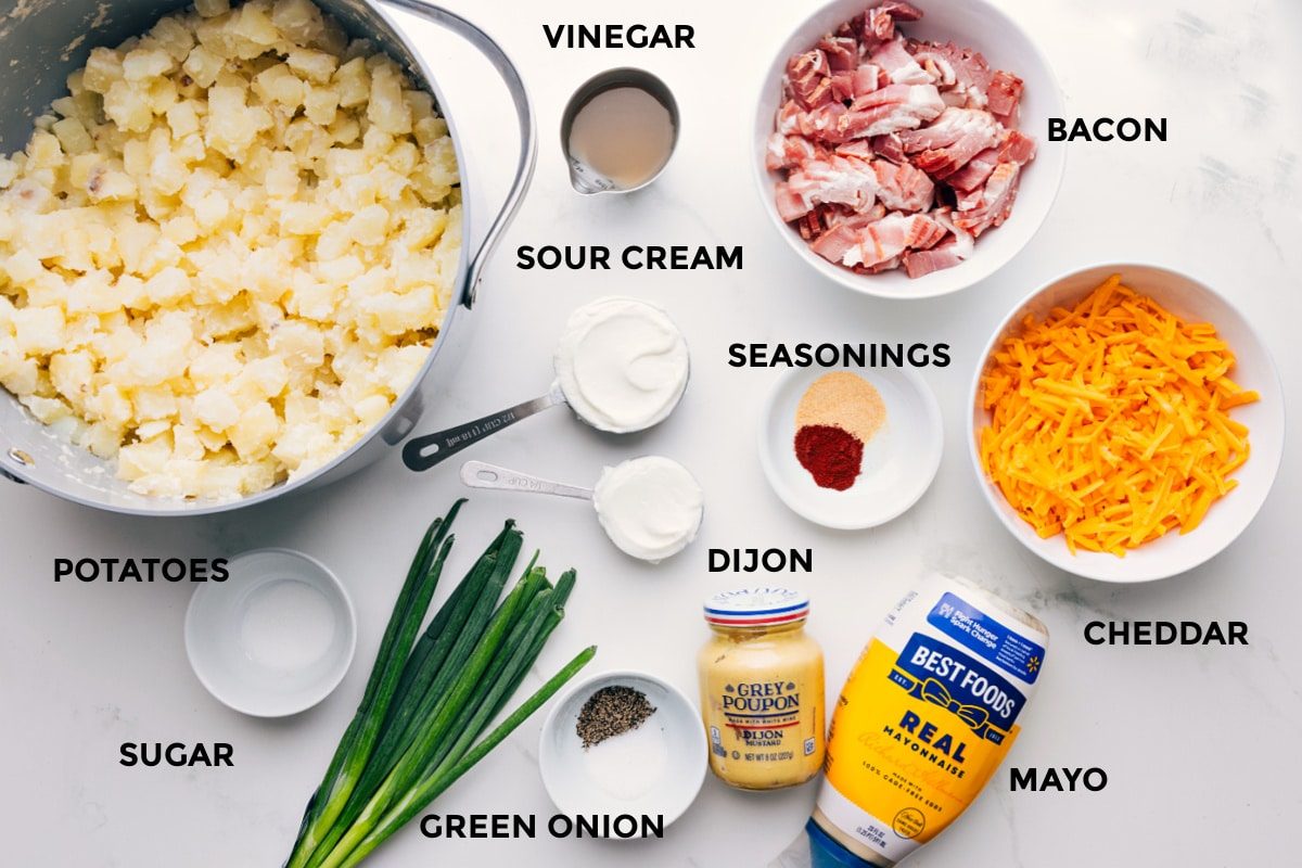 All the ingredients in this recipe prepped out for straightforward assembly including the potatoes, sugar, green onions, mayo, cheddar, seasonings, sour cream, vinegar, and bacon.