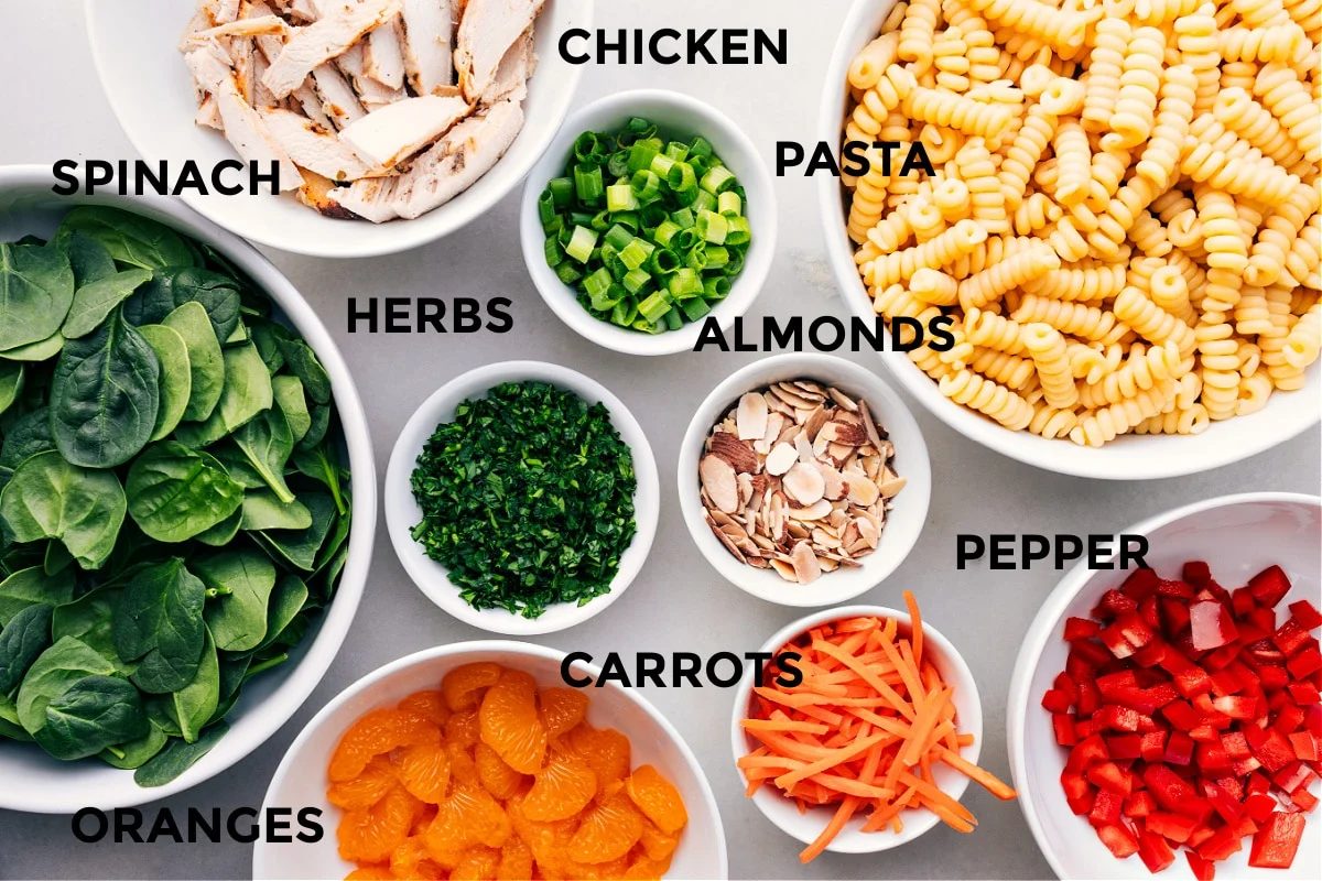 Asian pasta salad ingredients laid out for straightforward assembly including chicken, pasta, almonds, herbs, spinach, peppers, carrots, and oranges.