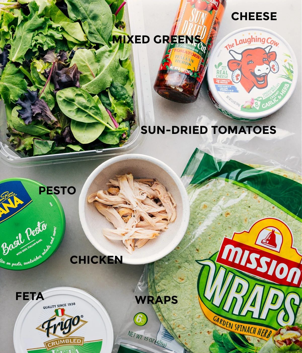 All the ingredients in this recipe prepped out for straightforward assembly, including pesto, chicken, mixed greens, cheese, tortillas, and feta.