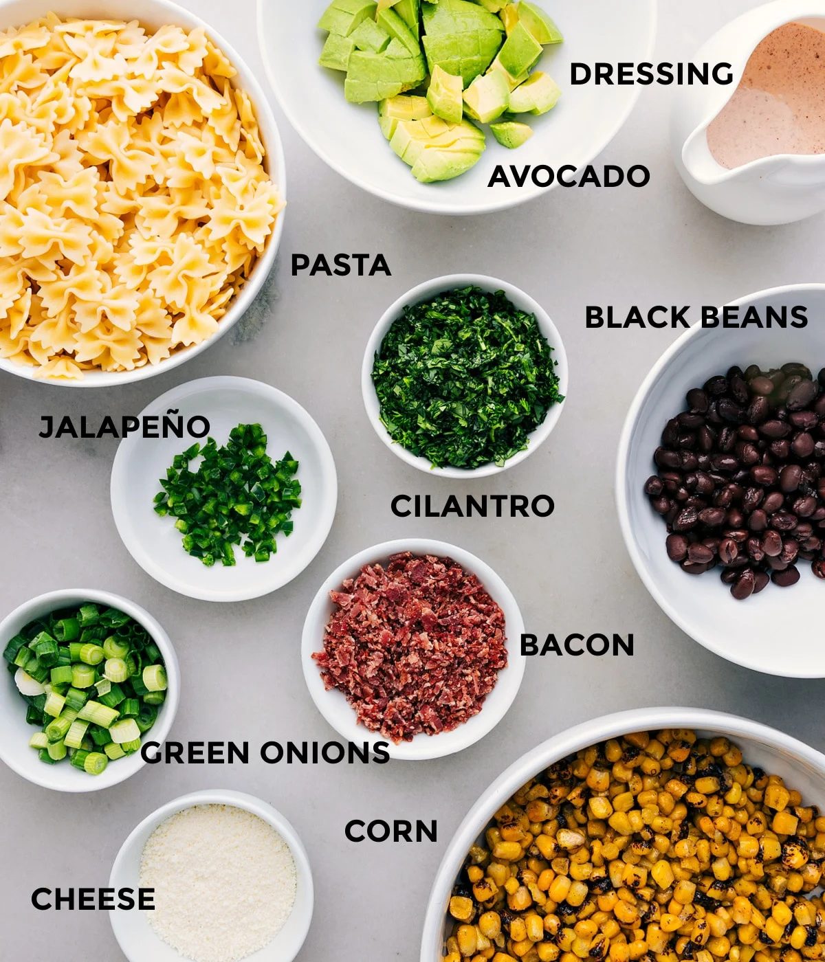 All the ingredients for this recipe prepped and ready for straightforward assembly: dressing, avocado, black beans, cilantro, noodles, bacon, green onions, corn, and cheese.