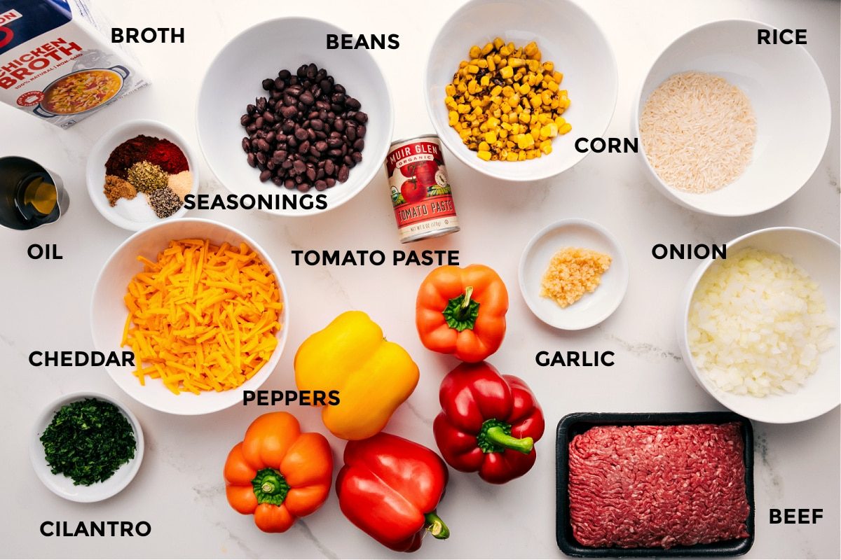 Stuffed pepper ingredients prepped for straightforward assembly, including broth, oil, spices, cheddar cheese, cilantro, peppers, beans, tomato paste, garlic, ground beef, rice, corn, and onions.