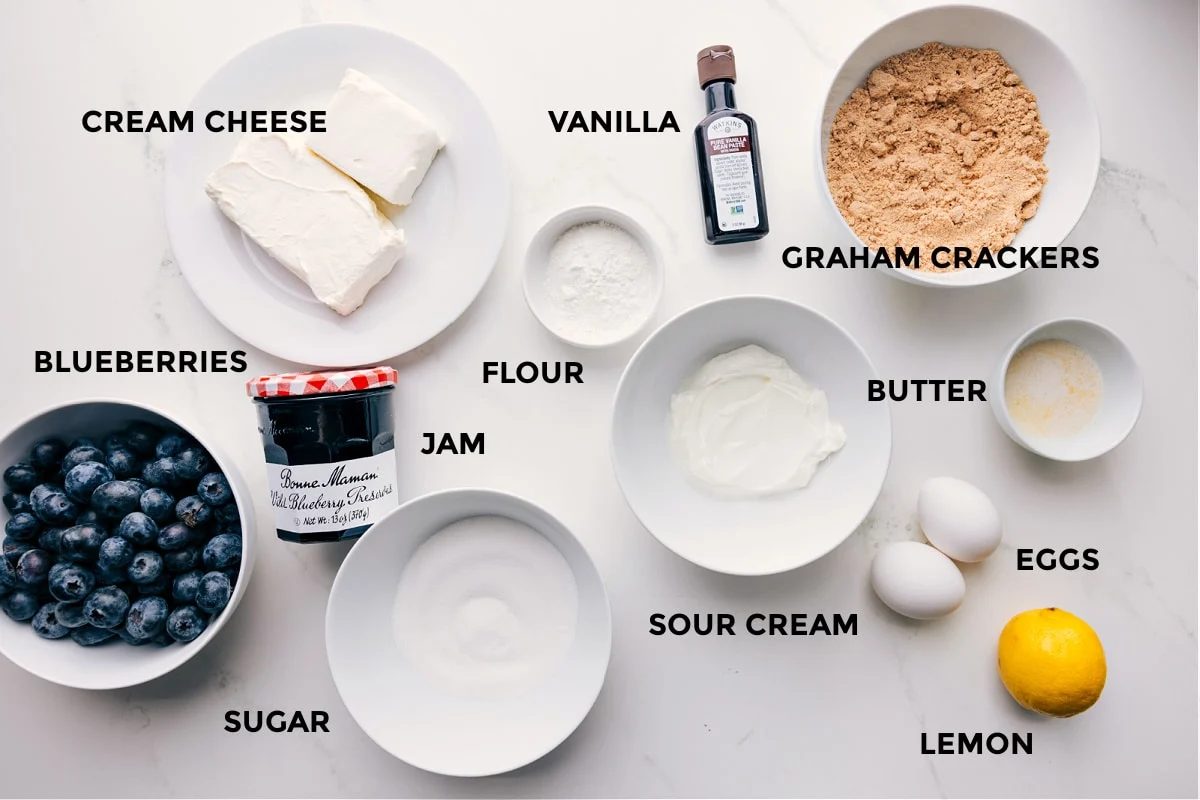 All the ingredients in this recipe prepped out for straightforward assembly including the cream cheese, blueberries, jam, sugar, flour, sour cream, eggs, lemon, butter, graham crackers, and vanilla.