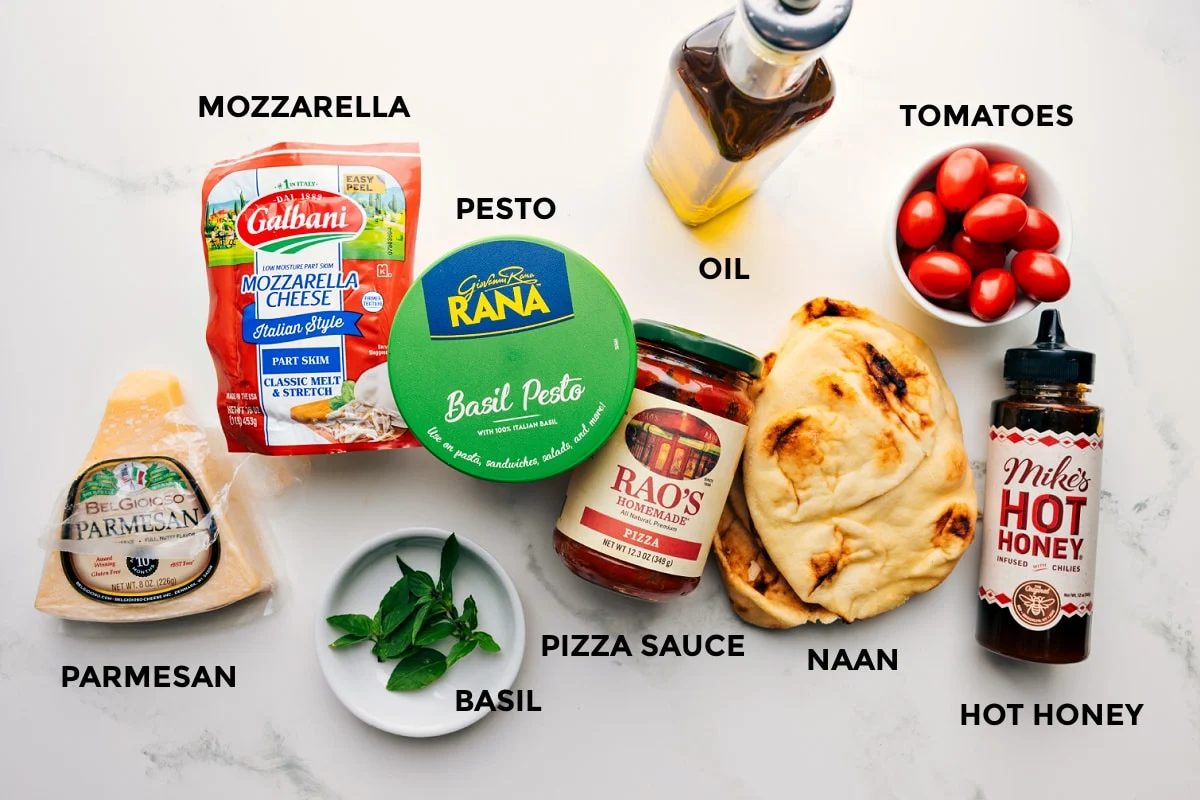 All the ingredients prepped out for straightforward assembly including the parmesan, mozzarella, pesto, basil, sauce, oil, naan, hot honey, and tomatoes.