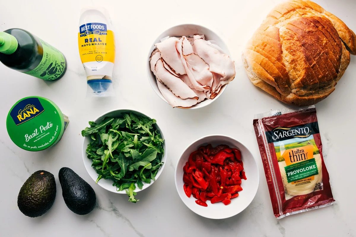 All the ingredients in this recipe prepped out for straightforward assembly including the oil, pesto, avocado, arugula, turkey, roasted red peppers, provolone, and sourdough.