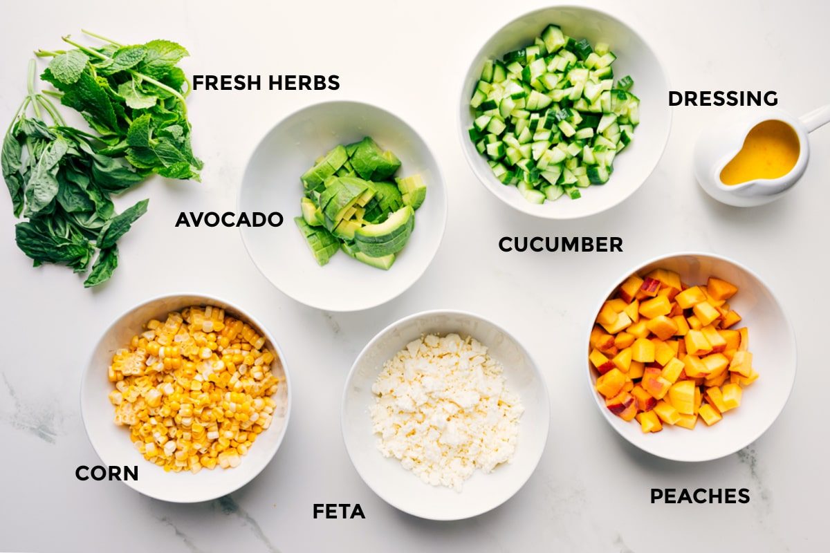 Ingredients in this recipe prepped out for straightforward assembly including the fresh herbs, avocado, corn, feta, cucumber, peaches, and dressing.