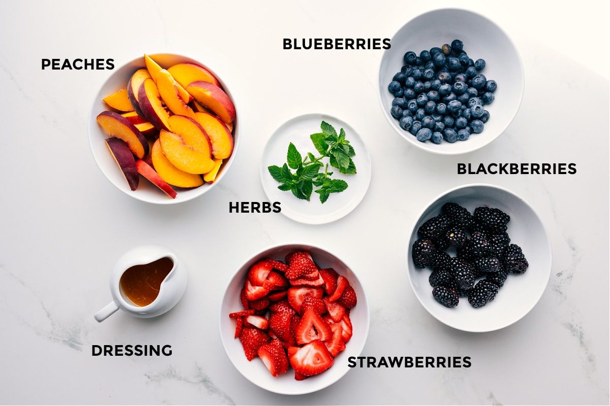 Ingredients in this recipe prepped out for straightforward assembly including peaches, herbs, blueberries, blackberries, strawberries, and dressing.