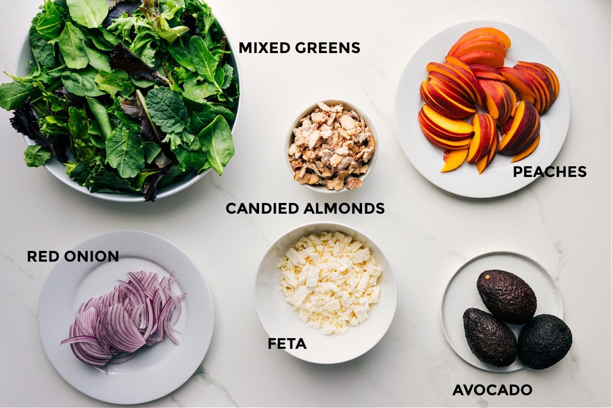 Ingredients in this recipe are prepped for straightforward assembly: mixed greens, red onions, feta, candied almonds, avocado, and peaches.