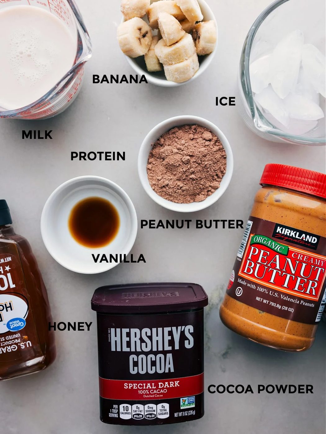 All the ingredients including cocoa powder, vanilla, peanut butter, protein powder, bananas, milk, ice, and honey prepped out for straightforward assembly.