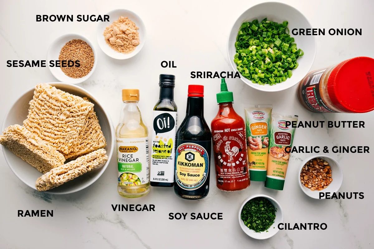 All the ingredients in this recipe prepped out for straightforward assembly including the ramen, sesame seeds, brown sugar, vinegar, oil, soy sauce, sriracha, cilantro, peanut butter, garlic, ginger, peanuts, and green onions.