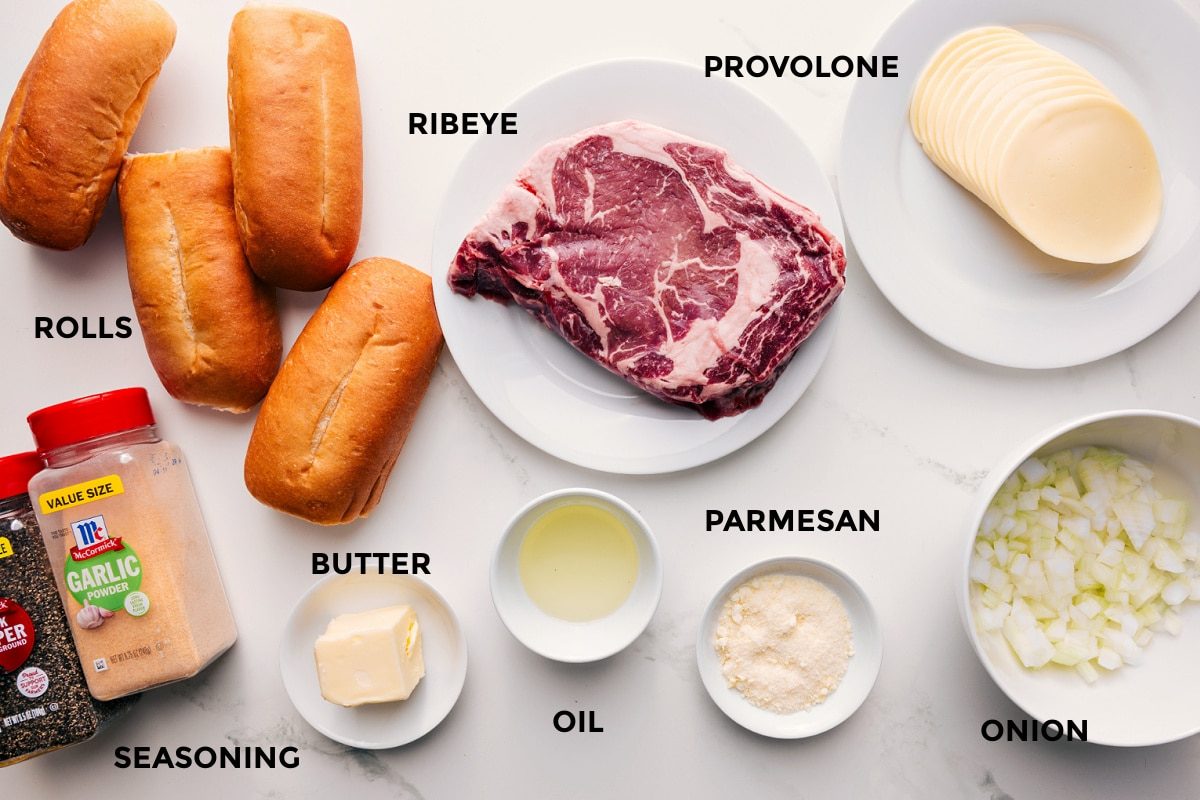 All the ingredients in this recipe prepped out for straightforward assembly including the rolls, seasonings, butter, oil, parmesan, ribeye, provolone, and onions.