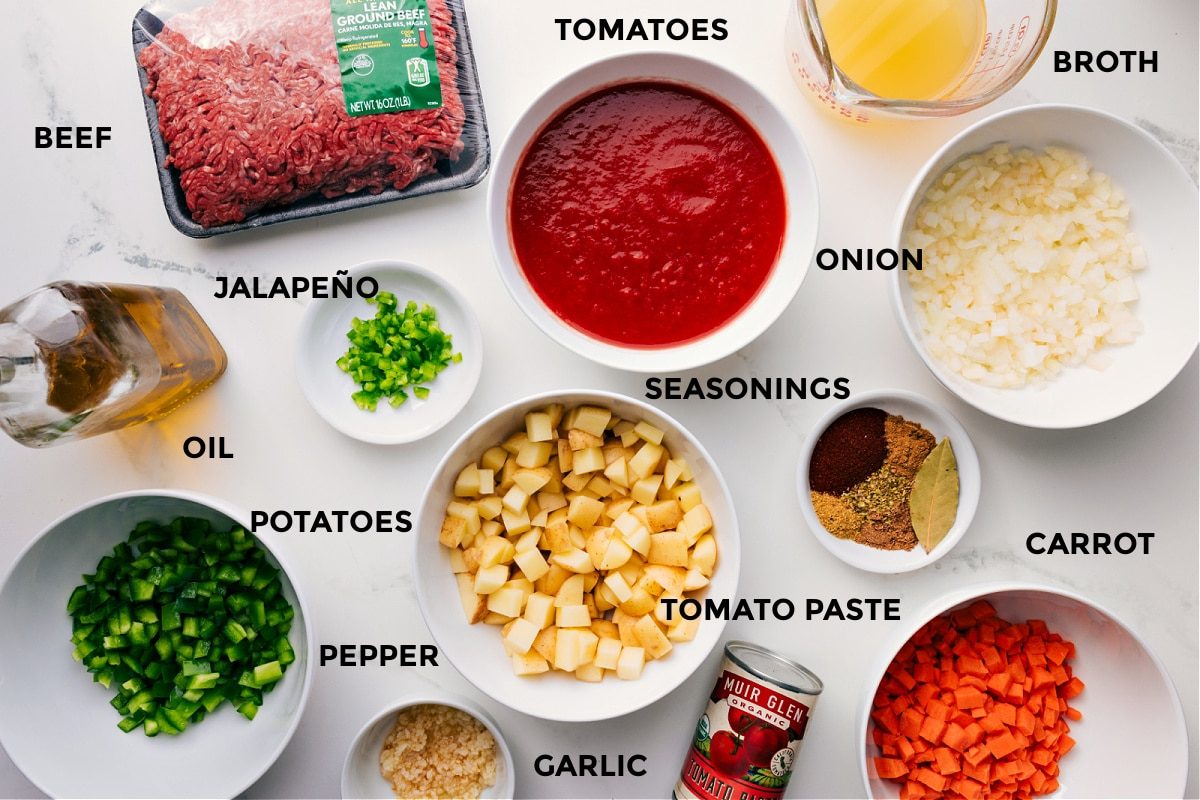 All the ingredients for this recipe prepped and ready for straightforward assembly, including the beef, broth, tomatoes, onions, seasonings, carrot, tomato paste, potatoes, jalapeño, oil, pepper, and garlic.