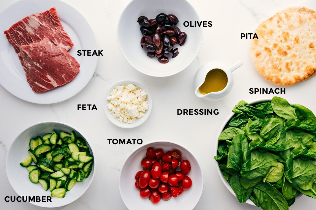 All the ingredients prepped for straightforward assembly, including sliced steak, olives, pita wedges, spinach, honey-lemon dressing, crumbled feta, cherry tomatoes, and cucumber.