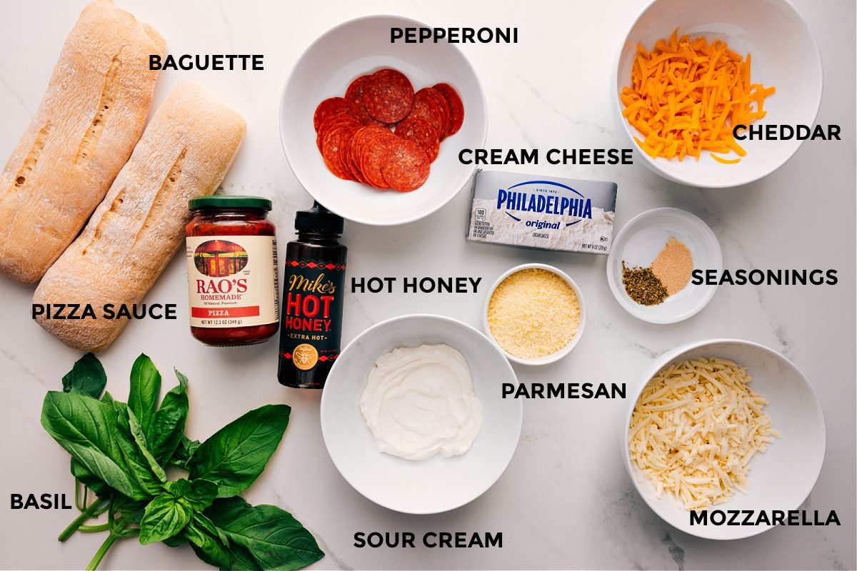 All the ingredients in this recipe prepped out for straightforward assembly including the baguette, basil, pizza sauce, hot honey, sour cream, pepperoni, cream cheese, parmesan, mozzarella, seasonings, and cheddar.