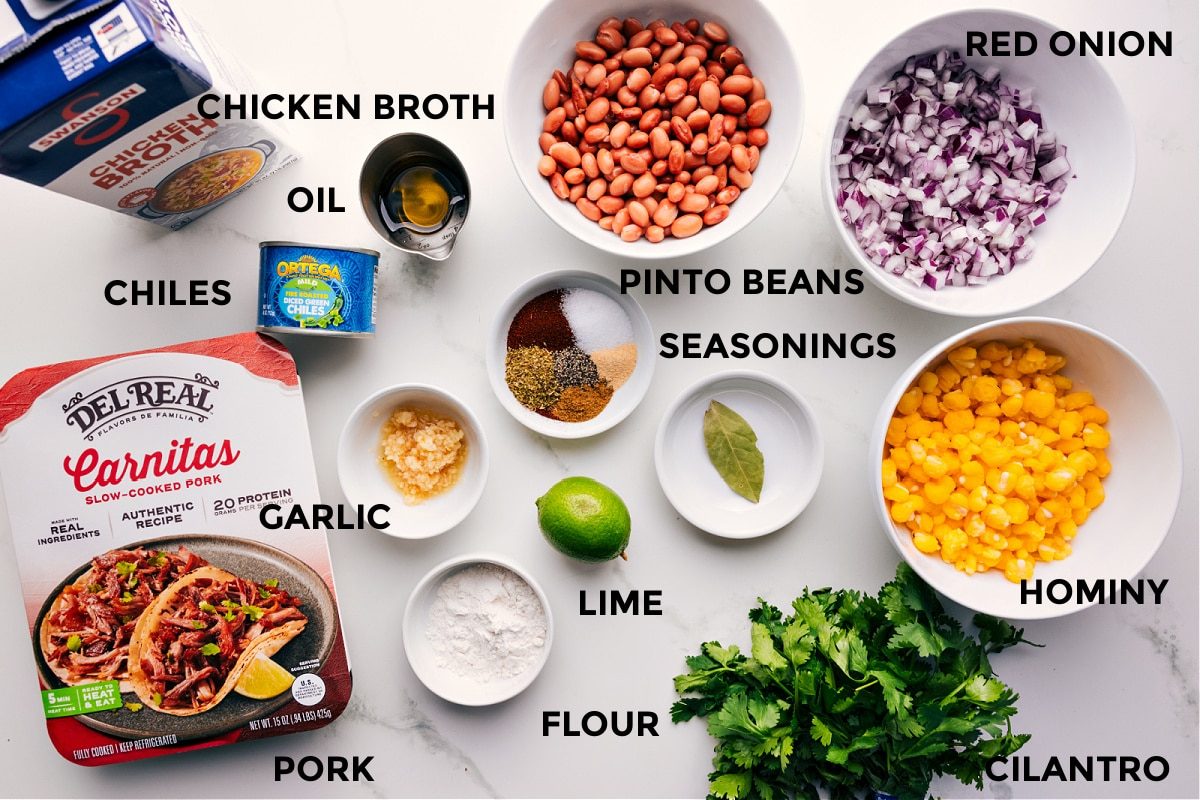 All the ingredients for this recipe prepped and ready for straightforward assembly, including broth, oil, chiles, garlic, pork, flour, limes, pinto beans, seasonings, hominy, cilantro, and red onions.