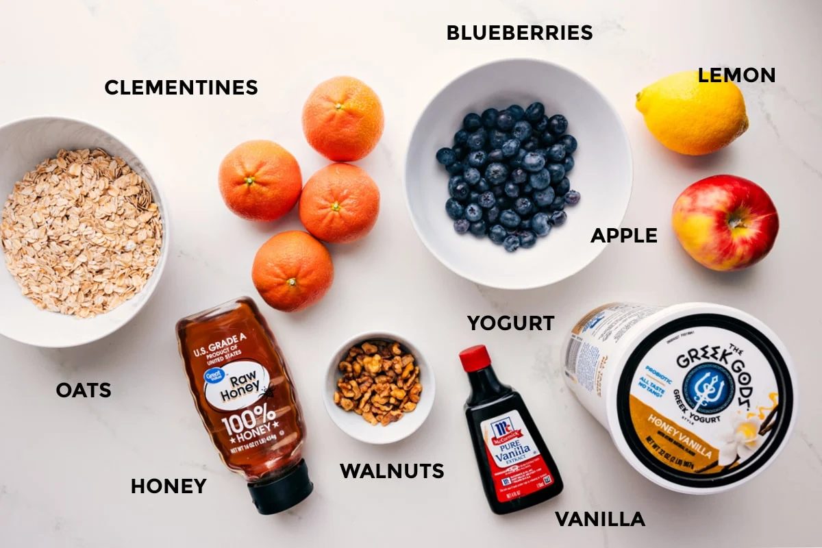 Ingredients in this recipe prepped out for straightforward assembly including the oats, honey, walnuts, clementines, blueberries, yogurt, vanilla, lemon, and apple.
