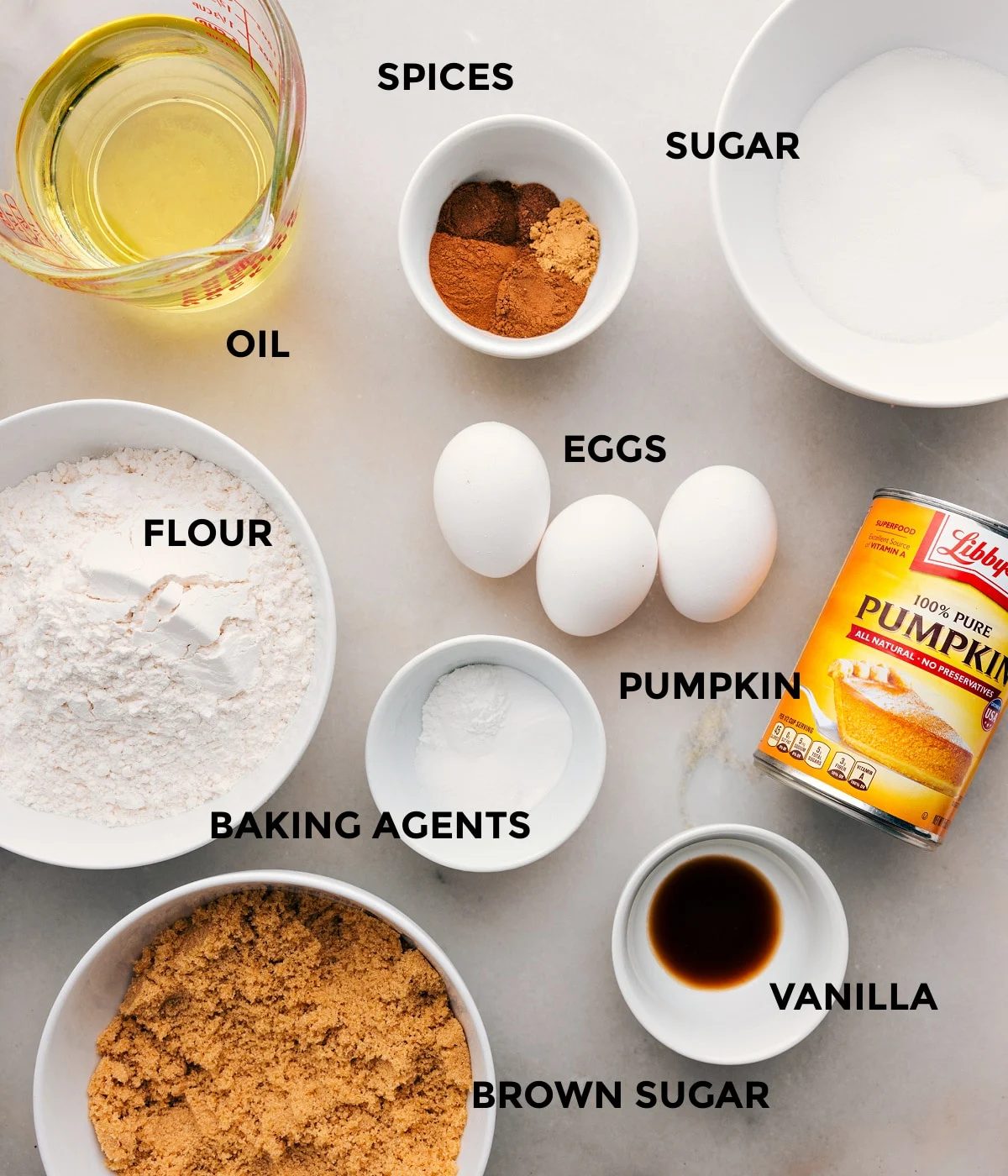 All the ingredients in this recipe including oil, sugar, spices, eggs, flour, baking agents, and pumpkin prepped out for straightforward assembly.