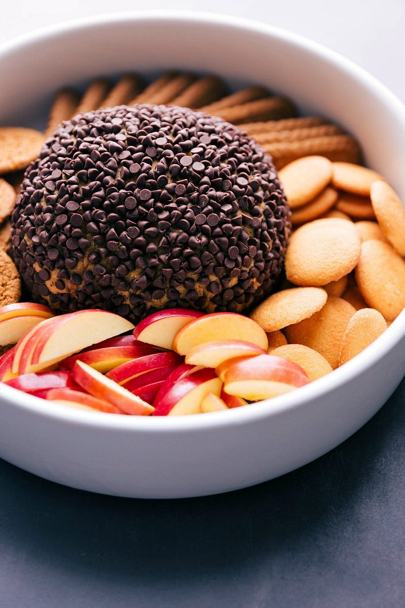 A satisfying no bake pumpkin cheesecake ball presented in a bowl, surrounded by an assortment of dippers including vanilla wafers, apple slices, and more, offering a delightful and indulgent dessert spread.
