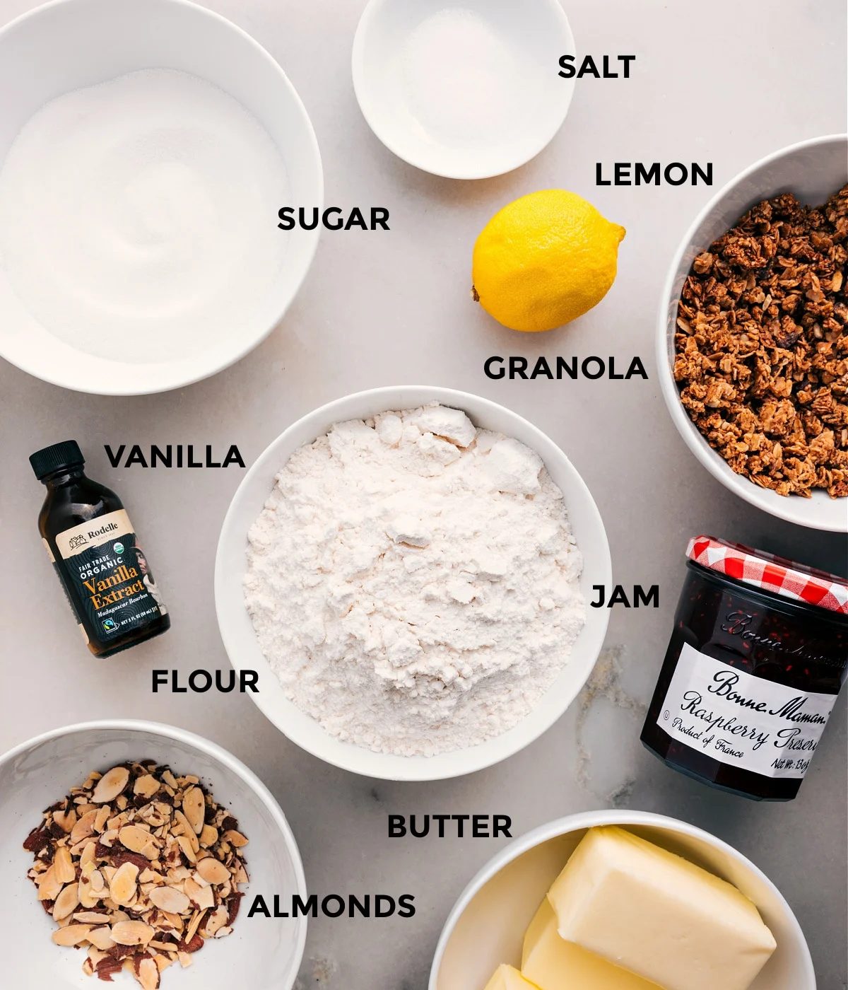 All the ingredients prepped and ready for straightforward assembly—lemon, granola, jam, flour, vanilla, sugar, salt, butter, and almonds.