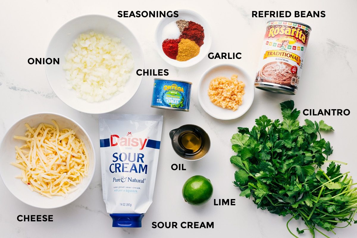 All the ingredients in this recipe prepped out for straightforward assembly including the cheese, onion, sour cream, lime, oil, chiles, seasoning, garlic, refried beans, and cilantro.