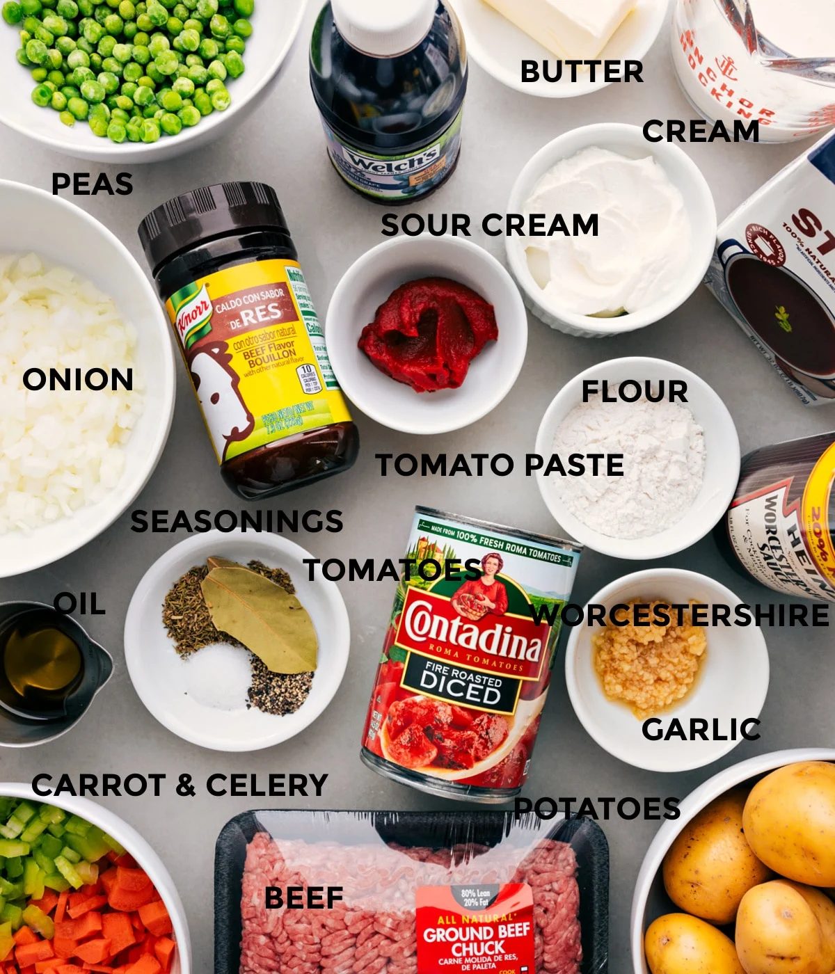 All the ingredients in this recipe prepped out for straightforward assembly including the peas, onions, seasonings, tomatoes, sour cream, tomato paste, grape juice, butter, cream, stock, flour, Worcestershire, garlic, potatoes, and beef.