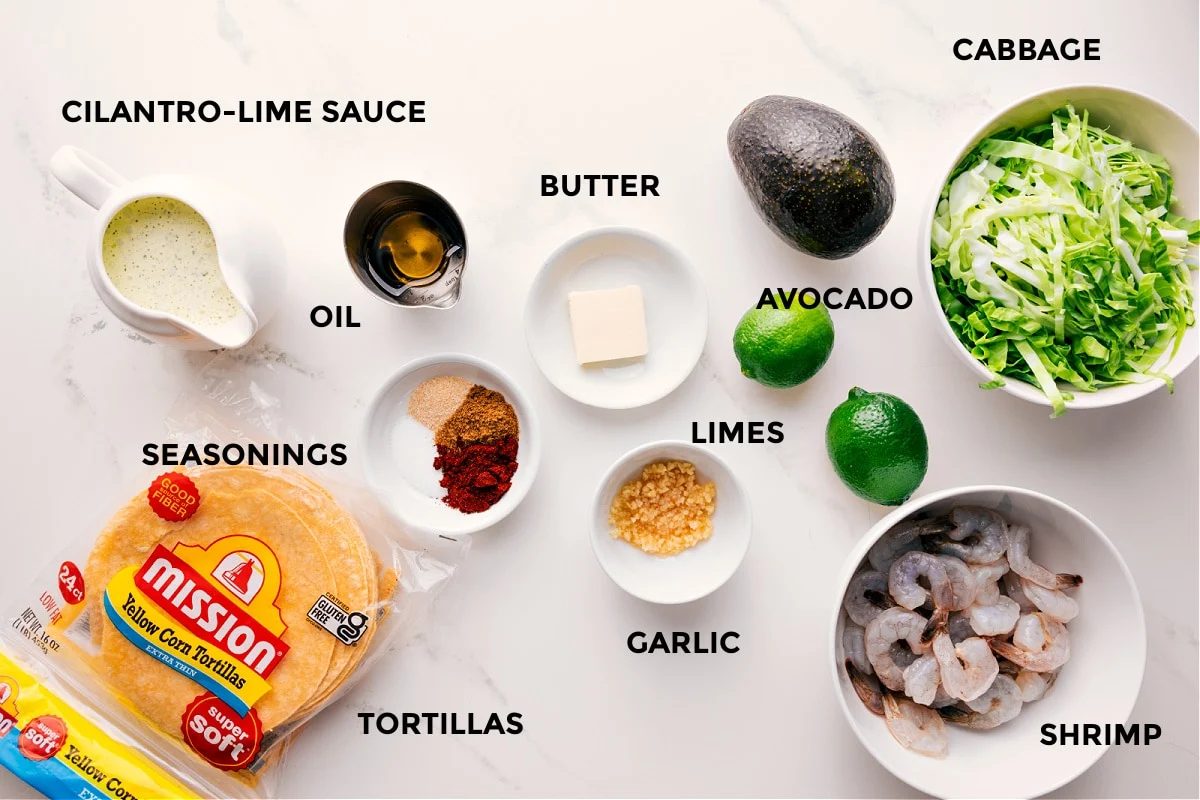 All the ingredients in this recipe prepped out for straightforward assembly including the cilantro lime sauce, seasonings, tortillas, butter, garlic, limes, avocado, shrimp, and cabbage.